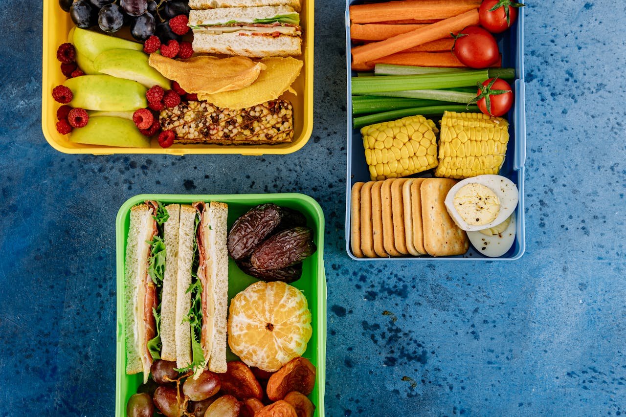Three vibrant lunch boxes filled with a variety of healthy snacks and sandwiches on a blue surface.